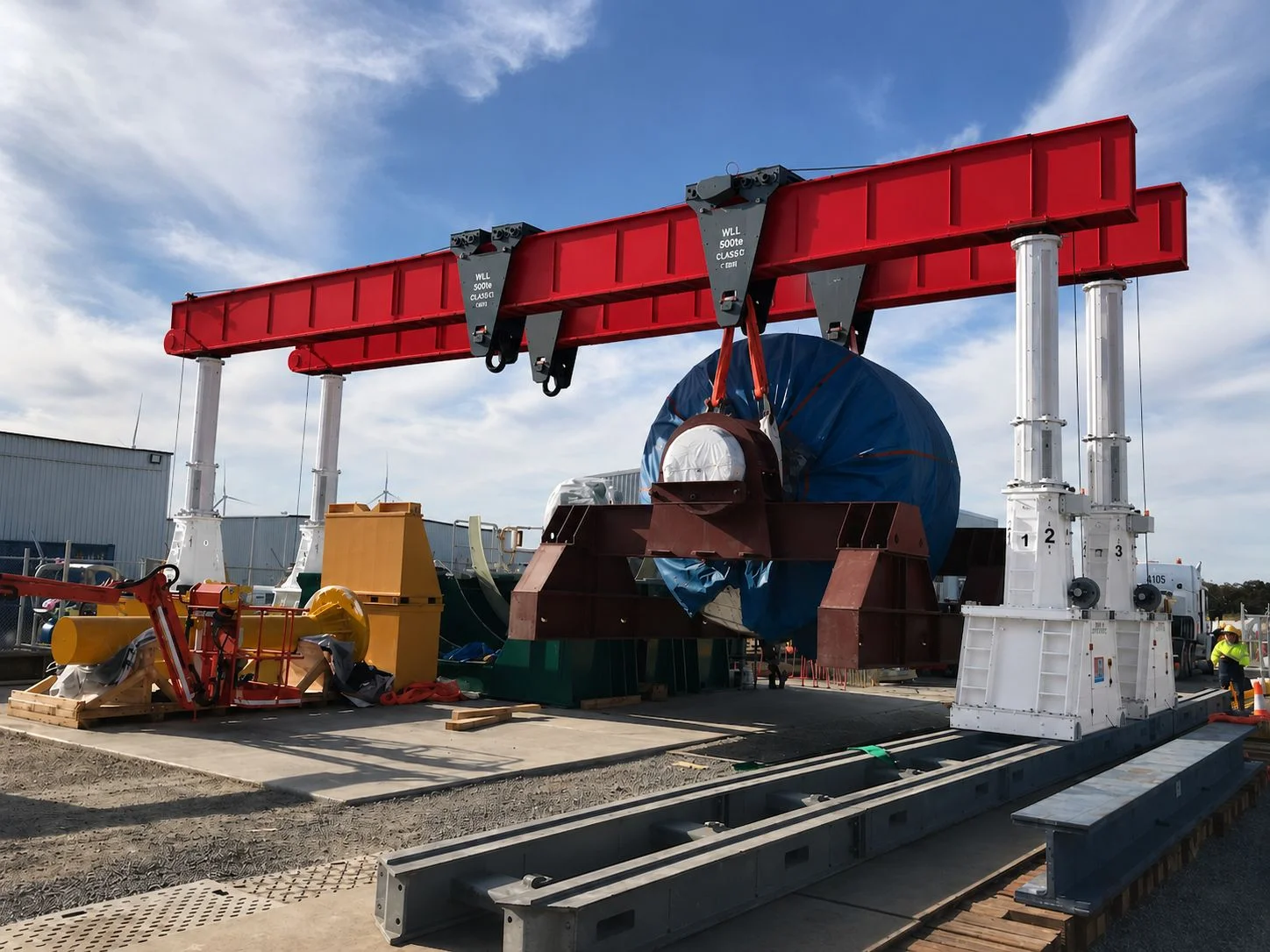 A large industrial gantry crane with red beams lifts a massive cylindrical object wrapped in blue material at an outdoor construction or industrial site under a blue sky. Workers and equipment are visible nearby.