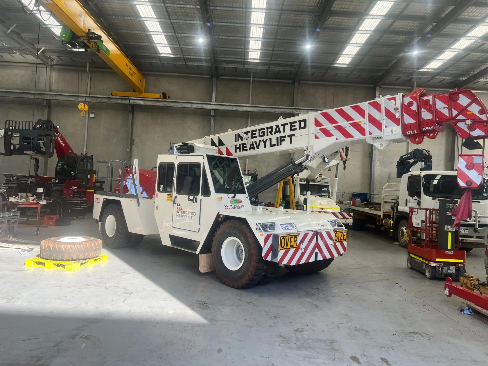 A large white crane truck with red stripes labeled "Integrated Heavylift" is parked inside an industrial warehouse, surrounded by machinery, tools, and spare tires.