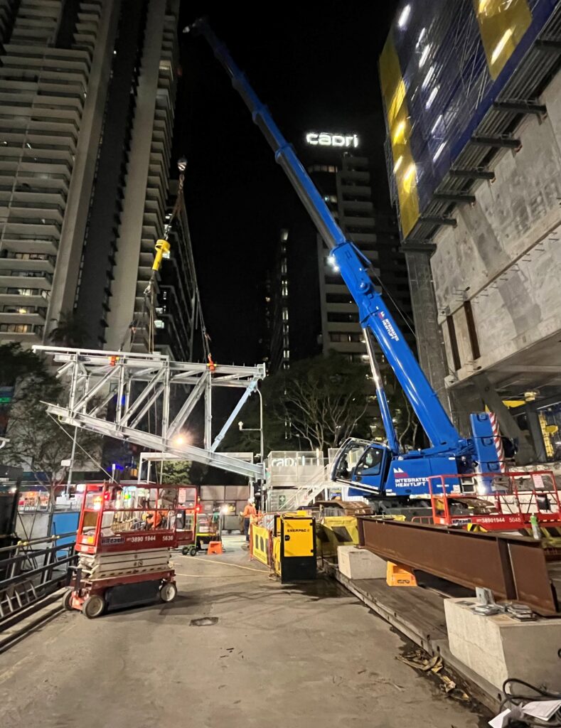 A blue crane lifts a large steel structure at a nighttime construction site between tall buildings, with workers, equipment, and illuminated signs visible in the background.