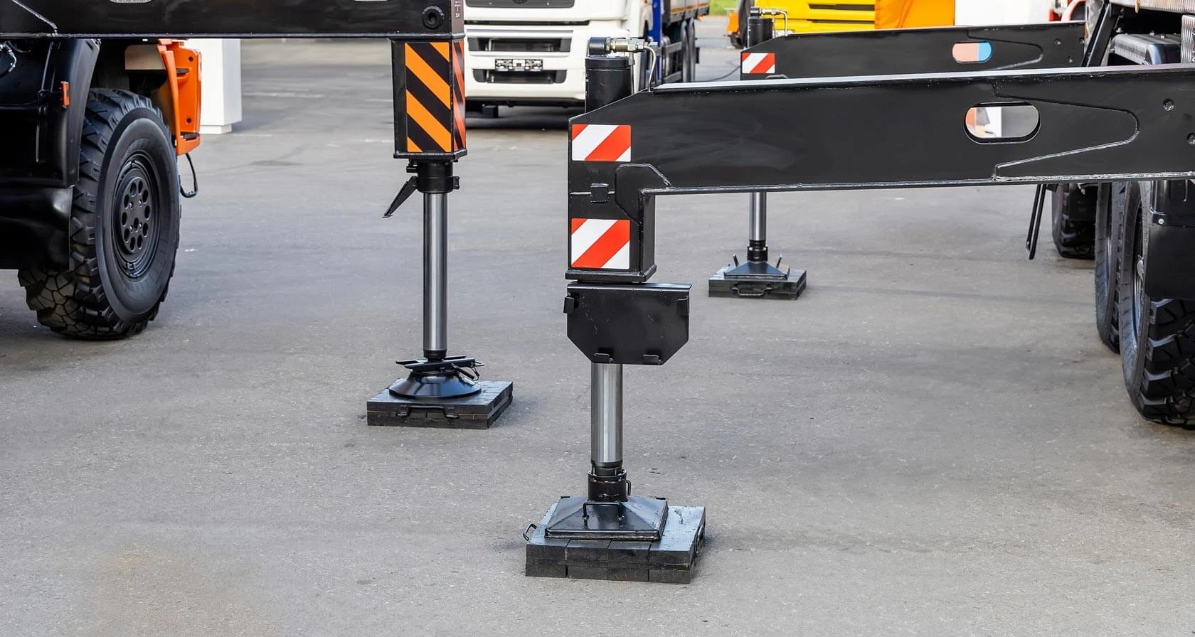 Hydraulic stabilizers with black and orange warning stripes extend from heavy vehicles, supporting the machinery on a level asphalt surface in an industrial or construction equipment setting.