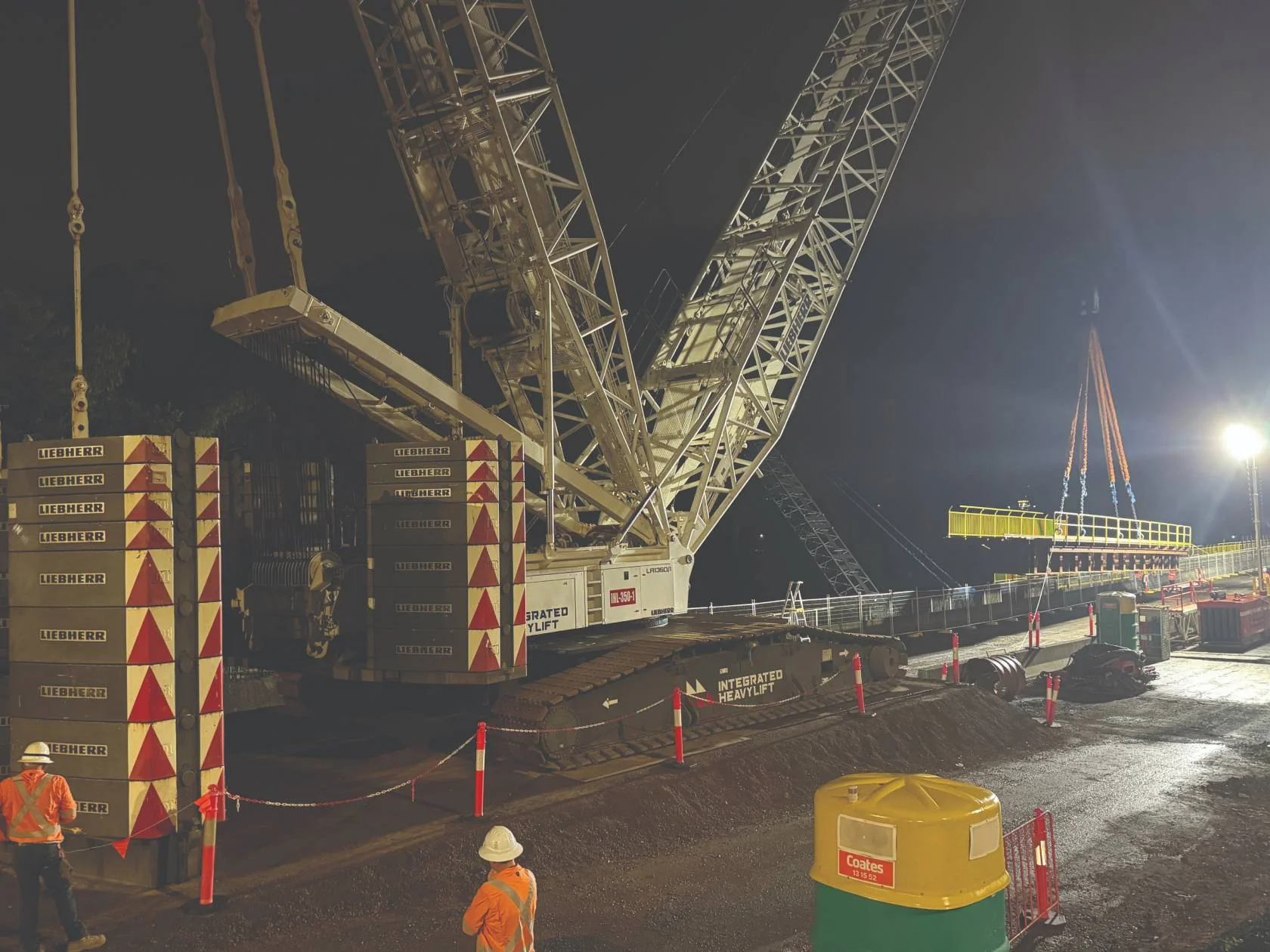 A large crane operates at a construction site at night, illuminated by bright floodlights. Workers in orange safety vests and helmets stand nearby, and construction barriers and equipment are visible.