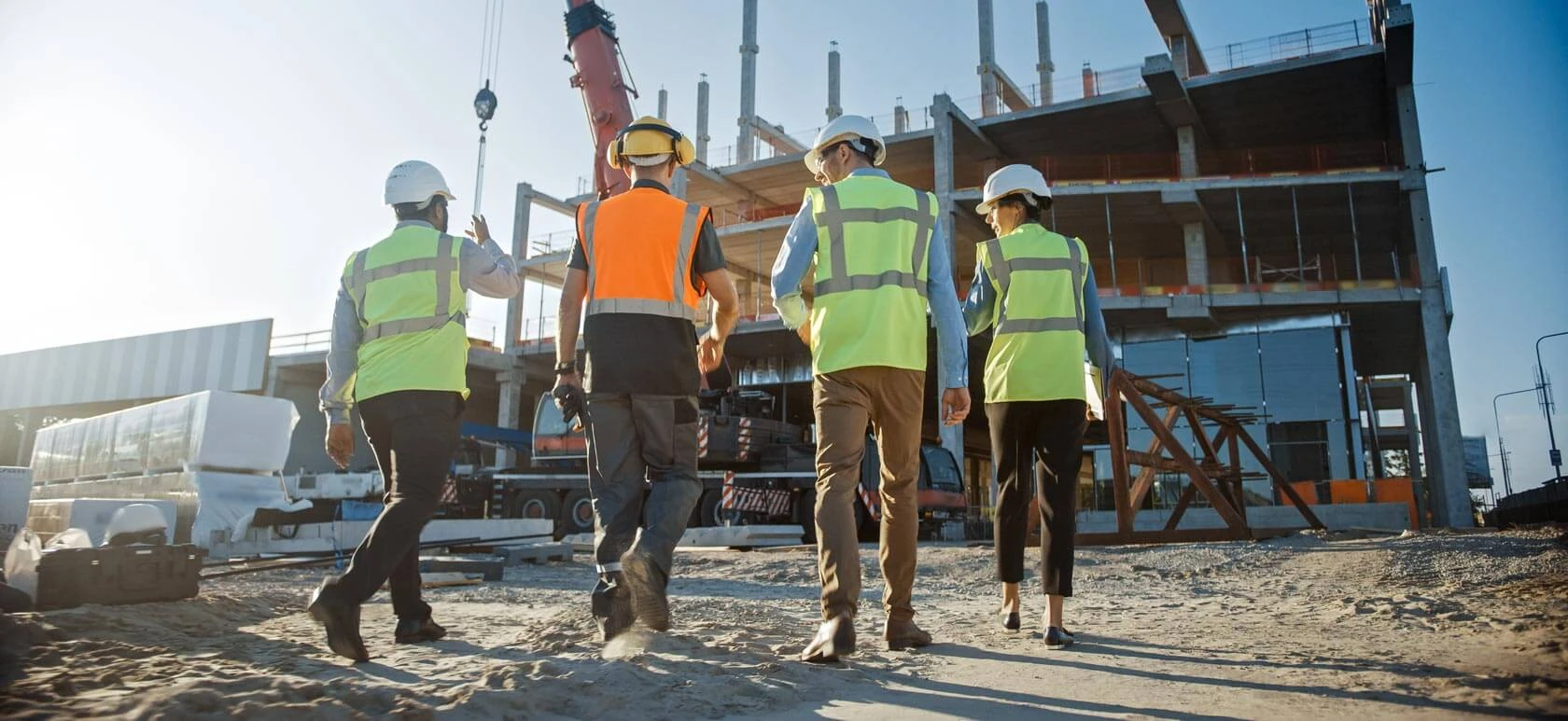 Four construction workers wearing safety vests and helmets walk toward a building under construction, with scaffolding, a crane, and construction materials visible in the background under a clear sky.