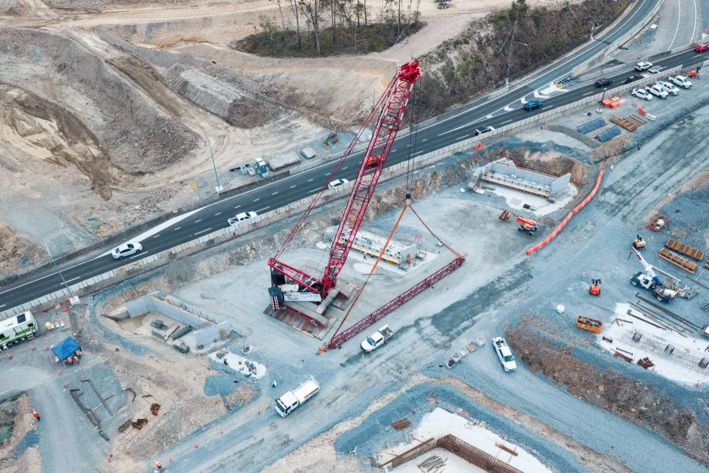 Aerial view of a construction site beside a roadway, featuring a large red crane, construction vehicles, and building materials. Traffic moves along the adjacent road, with some cleared and excavated land nearby.
