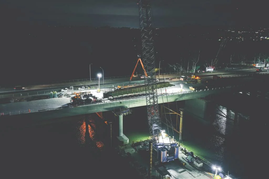 A nighttime construction scene shows workers and machinery on a partially built bridge over a body of water, illuminated by bright floodlights. A large crane is positioned on the bridge deck.