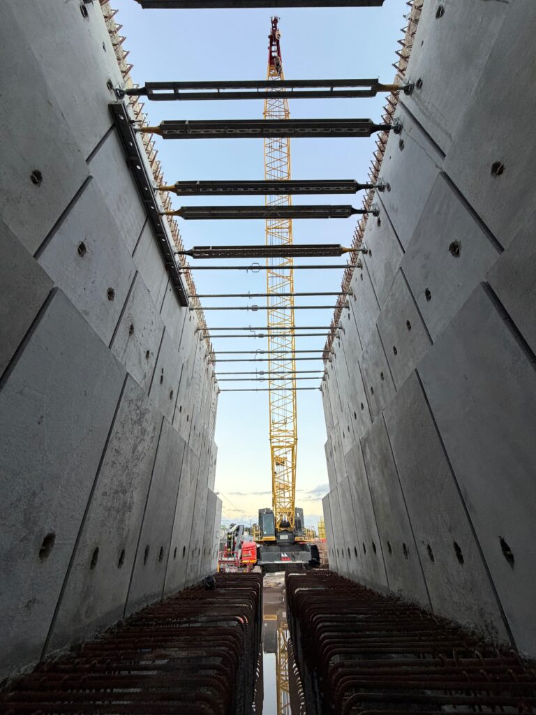 View between two concrete walls with exposed rebar at the bottom, looking toward a tall yellow construction crane in the background, under a blue sky with scattered clouds.