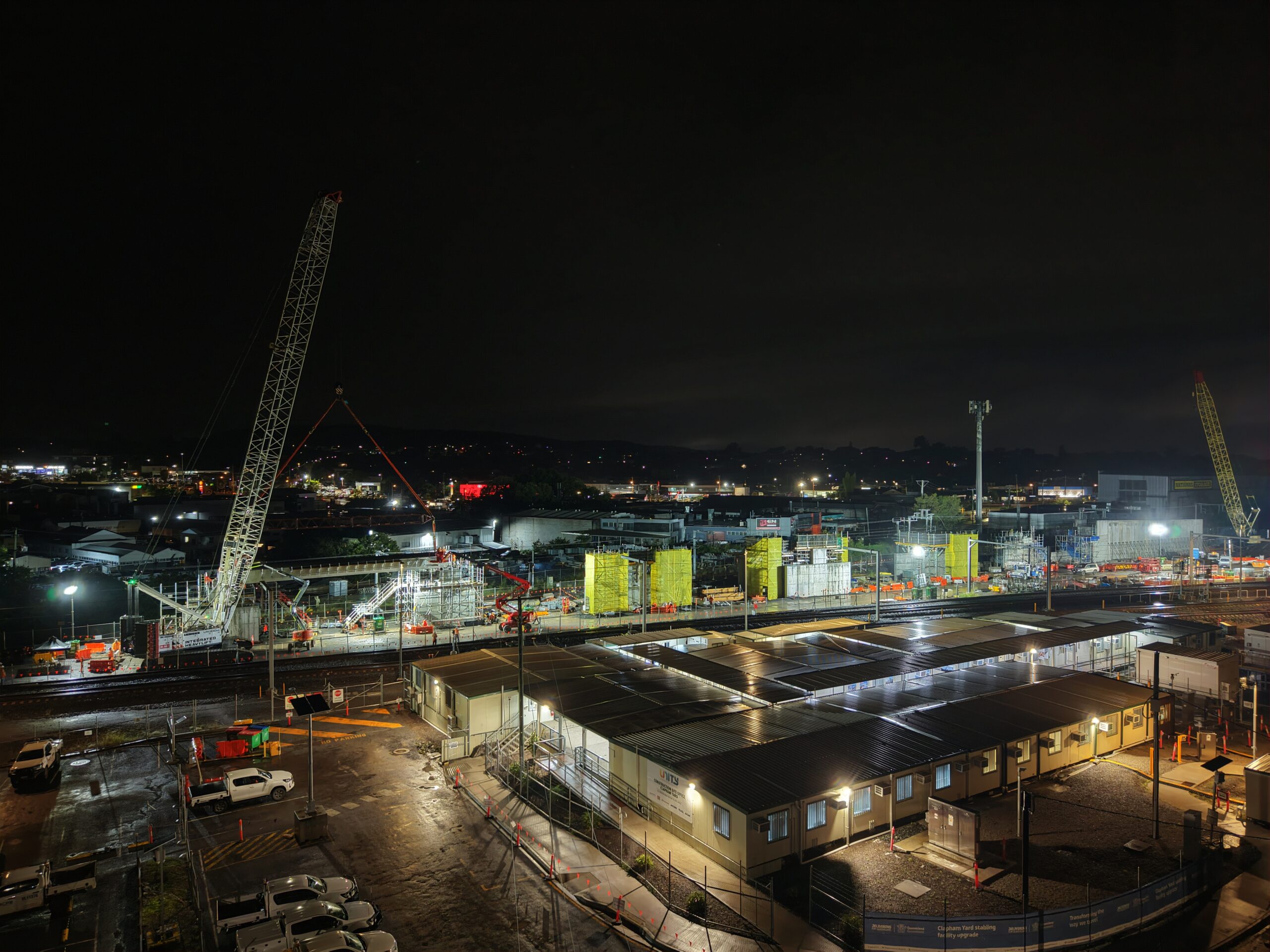 Nighttime view of a large construction site with cranes, bright floodlights, and several temporary buildings in the foreground, surrounded by parked vehicles and city lights in the distance.