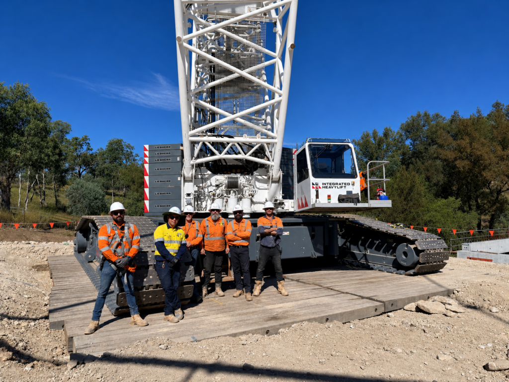 Five construction workers in safety gear stand in front of a large white crane on a construction site, with trees and a clear blue sky in the background.