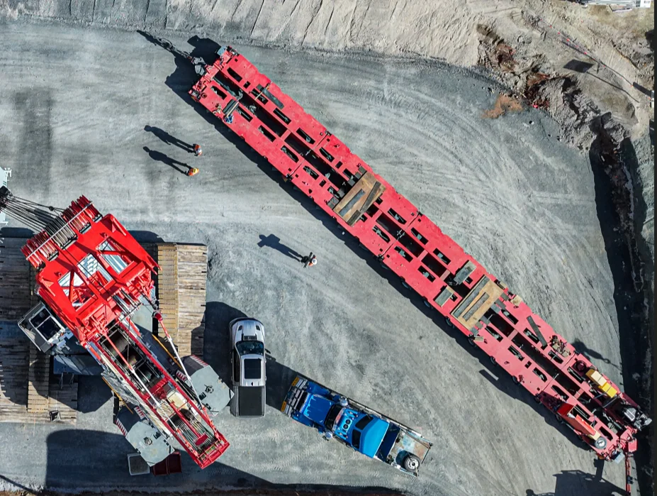 Aerial view of a construction site with a large red crane and a long red steel structure on gravel, surrounded by trucks, cars, wooden platforms, and several workers visible on the ground.