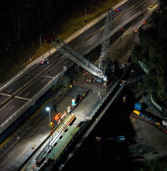 Aerial view of a nighttime construction site beside a road with a large crane, workers, and vehicles illuminated by work lights, while cars drive on the adjacent highway.
