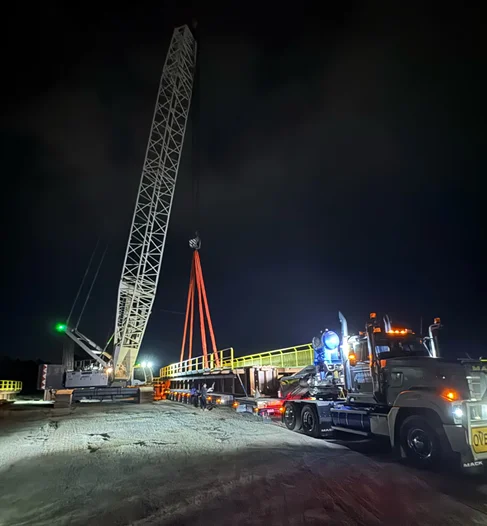 A large crane lifts a long metal structure at a construction site at night. A truck with flashing lights is parked nearby, and the area is illuminated by work lights.