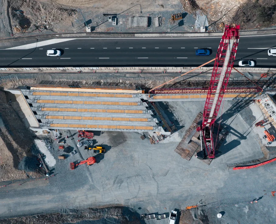 Aerial view of a construction site with a large red crane and several vehicles near a partially built bridge, adjacent to a road with cars driving on it. Gravel and construction equipment are visible around the site.