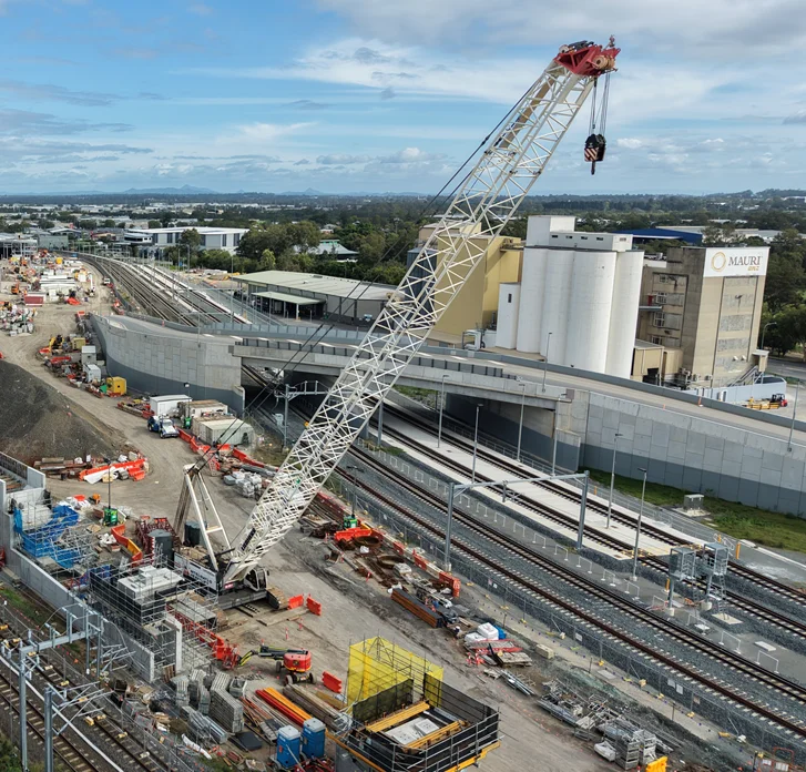 A large crane operates on a construction site next to multiple railway tracks. Nearby are construction materials, equipment, and workers, with buildings and roads in the background under a partly cloudy sky.
