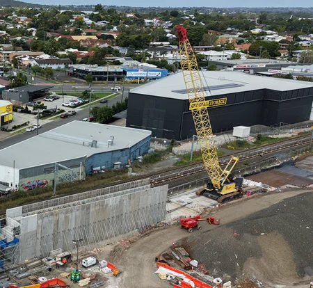 A yellow crane operates next to a large retaining wall under construction beside train tracks, with nearby storage buildings and a suburban neighborhood in the background. Construction equipment is scattered on the site.