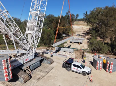 A large crane lifts a bridge section over a dry, dirt construction site, while workers and a white utility vehicle are present. Trees and a partly cleared hill are visible in the background.