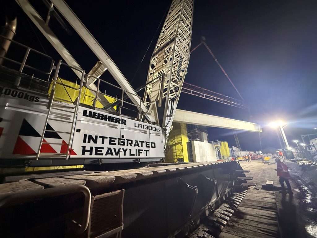 A large Liebherr LR1300SX crane with "INTEGRATED HEAVYLIFT" branding lifts a steel beam at a construction site at night, illuminated by bright lights. Workers in safety gear stand nearby.