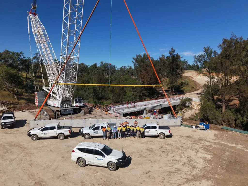 A group of construction workers stand in front of several white vehicles and a large crane at a bridge construction site, surrounded by trees and dirt under a clear blue sky.